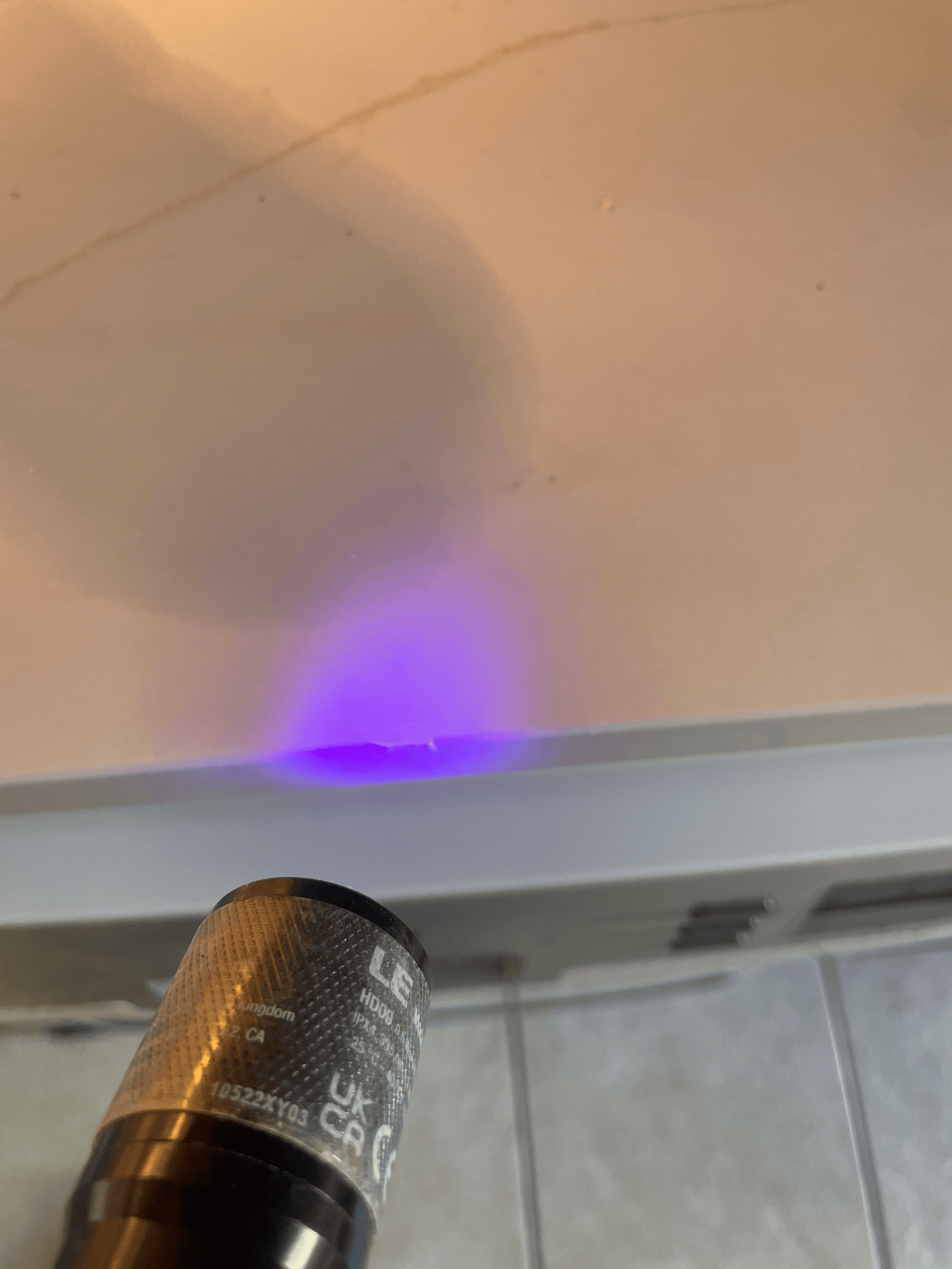 Handheld UV flashlight emitting a purple glow onto a white wall in a close-up.
